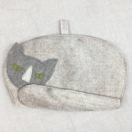 A photo of the cat shaped tea cosy laid flat against a smooth linen cloth. The cat has a pale grey body and dark grey head with green eyes. 